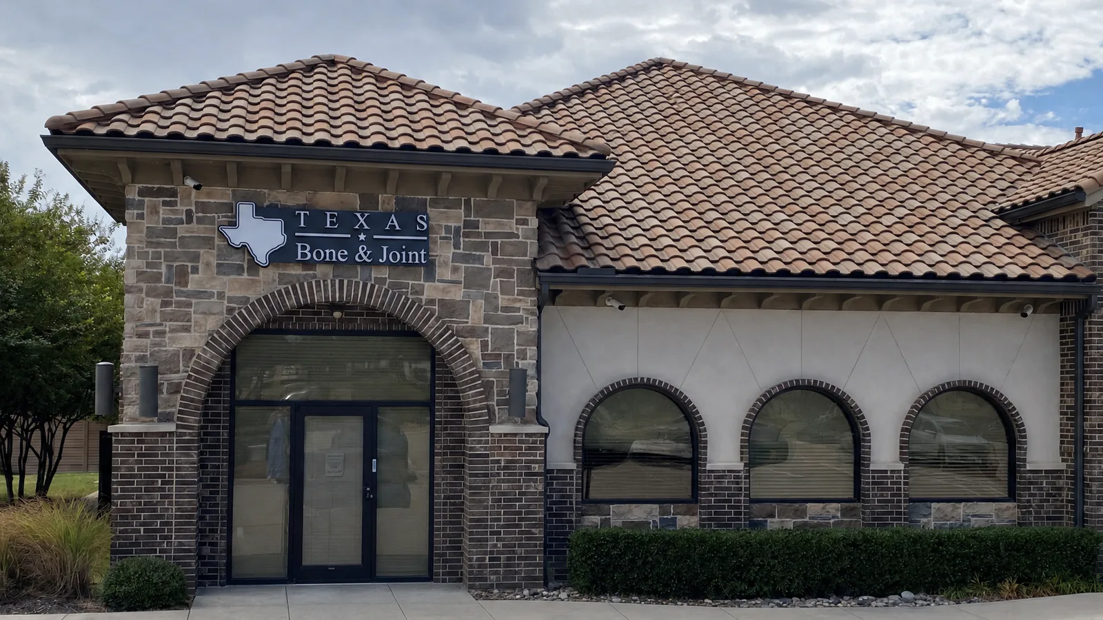 The Texas Bone and Joint clinic in Keller: a stone and brick building with a clay tile roof, three arched windows, and the practice sign above an arched entrance